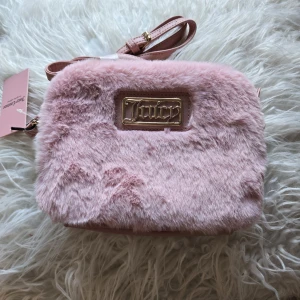 Rosa fluffig axelväska från Juicy Couture - Supersöt axelväska från Juicy Couture i ljusrosa fuskpäls med guldig logga framtill. Väskan har justerbar axelrem i rosa syntetmaterial och dragkedja upptill. Insidan är fodrad med Juicy Couture-tryck. Perfekt för dig som vill sticka ut med en mjuk och trendig accessoar.