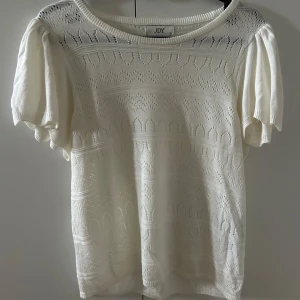 Vit stickad blus från JDY - Supersöt vit stickad blus från JDY med kort puffärm och rund halsringning. Blusen har ett genombrutet mönster och är gjord i ett mjukt stickat material som känns luftigt och fräscht. Perfekt att styla med jeans eller kjol för en chill look.
