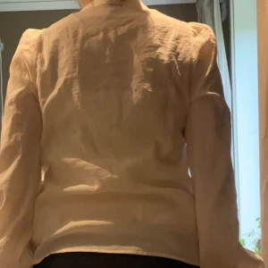 Beige blus med puffärm och knappar - Säljer en stilren beige blus i linne med v-ringning och knappar framtill. Blusen har långa puffärmar och en lätt oversized passform. Perfekt för dig som gillar en clean och luftig look. Snygg att styla med både jeans och kjol.