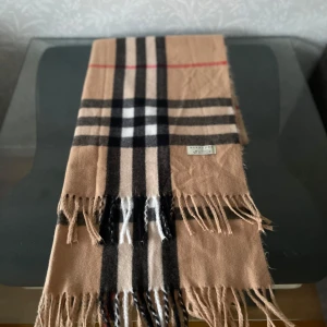 Beige rutig halsduk från Burberry - Snygg beige halsduk från Burberry i klassiskt rutigt mönster med svarta, vita och röda detaljer. Gjord i 100% kashmir, supermjuk och lyxig. Fransar i kanterna ger en chill vibe. Perfekt att slänga över axlarna när det är kallt ute. Priset kan diskuteras till lägre