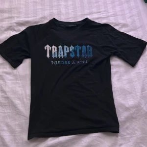 Svart Trapstar t-shirt med blått tryck - Svart t-shirt från Trapstar i storlek S med stort broderat Trapstar-tryck i vitt och blått på bröstet. Klassisk passform med rund hals och korta ärmar. Materialet är mjuk bomull som känns skönt mot huden. Perfekt för dig som gillar streetwear och vill sticka ut.