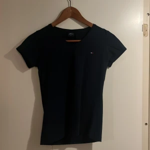 Svart t-shirt från Tommy Hilfiger - Klassisk svart t-shirt från Tommy Hilfiger med liten broderad logga på bröstet. T-shirten har v-ringning och korta ärmar, perfekt för en clean och stilren look. Tillverkad i mjuk bomull för maximal komfort.