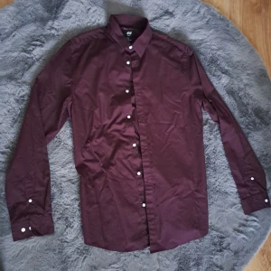 Vinröd skjorta slim fit från H&M - Vinröd skjorta från H&M i slim fit-modell med klassisk krage och vita knappar. Skjortan är långärmad och tillverkad i bomull, perfekt för dig som gillar en stilren och enkel look. Easy iron-tyg gör den smidig att hålla snygg.