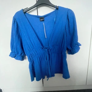 Blå blus med knyt från Gina Tricot - Supersöt blå blus från Gina Tricot i modellen Daisy top. Toppen har korta puffärmar, v-ringning och knyt i midjan. Materialet är strukturerat och ger en trendig look. Perfekt för dig som vill ha en färgklick i garderoben!