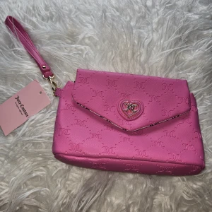 Rosa kuvertväska från Juicy Couture - Superfin rosa kuvertväska från Juicy Couture med JC-logga präglad över hela väskan och hjärtformad detalj framtill. Väskan har avtagbar handledsrem och fodrad insida med Juicy Couture-tryck. Perfekt för att lyfta din outfit med en poppig färg.