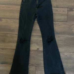 Svarta bootcut jeans med slitningar - Svarta jeans med bootcut passform och slitna hål på båda knäna. Jeansen har klassisk femficksdesign och är tillverkade i ett robust denimtyg. Perfekta för dig som gillar en edgy och avslappnad stil. Petite i längd.