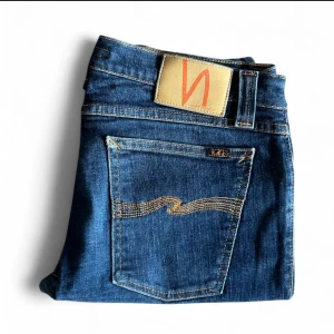 Blå jeans från Nudie Jeans Co - Snygga blå jeans från Nudie Jeans Co med klassisk femficksdesign och kontrastsömmar. Jeansen har rak passform och är tillverkade i slitstarkt bomullsjeans. Bakfickan har Nudie Jeans karaktäristiska broderi och läderpatch i midjan - W32 L32 - Hål vid höger insida lår (se bild) - Hör av er vid frågor eller funderingar