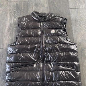 Moncler Gui Väst - Svart, glansig dunväst från Moncler med quiltad design och hög krage. Västen har två framfickor med dragkedja och Moncler-logga på bröstet. Materialet är syntet med dunfyllning, perfekt för att hålla värmen under kyliga dagar. Stängs med dragkedja framtill. Aldrig använd, helt ny. Säljer för den är för stor. 
