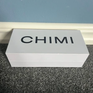CHIMI solglasögon transparent grå - Säljer ett par snygga CHIMI solglasögon med transparent grå båge och rektangulär form. Glasen är mörka och bågarna har en glittrig detalj med CHIMI-loggan på sidan. Kommer med originalfodral och box. Perfekt för dig som vill ha en clean och modern look.