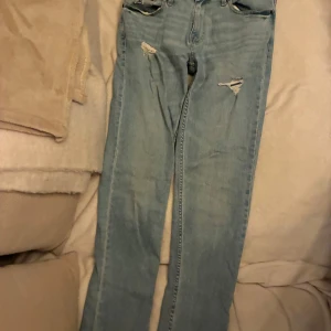 Ljusblå jeans med slitningar - Säljer ett par ljusblå skinny jeans med slitningar på låren. Jeansen har klassisk femficksdesign, bälteshällor och dragkedjegylf. Perfekta för dig som gillar en smal passform och en avslappnad streetstil.