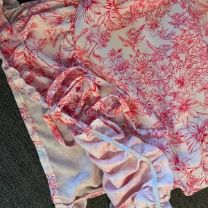 Rosa blommig kortklänning med volang - Supersöt kortklänning i vitt med rosa blommigt mönster. Klänningen har smala axelband, volangdetaljer och snörning i sidan. Perfekt för sommarens alla tillfällen och ger en fräsch och romantisk vibe.