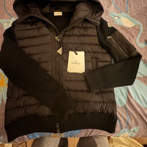 Svart dunväst med huva från Moncler - Säljer en svart dunväst från Moncler med huva och ribbade detaljer. Västen har quiltad framsida, dragkedja och en ficka på ärmen med Moncler-logga. Perfekt för lager-på-lager och snygg till streetwear.Pris kan diskuteras om det rimligt!!