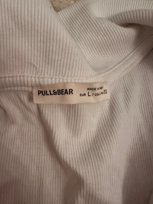 Vit ribbad polotopp från Pull&Bear - Säljer en vit ribbad polotopp från Pull&Bear i storlek L. Toppen har korta ärmar, krage och en snygg v-ringad öppning framtill. Materialet är mjukt och stretchigt, perfekt för en chill och stilren look.
