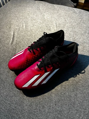 Adidas rosa och svarta fotbollsskor - Säljer ett par snygga Adidas fotbollsskor i rött och svart med vita ränder på sidorna. Skorna har snörning och en lätt, syntetisk ovandel samt mönstrad sula med dobbar för bra grepp på planen. Perfekta för dig som vill sticka ut på matchen!