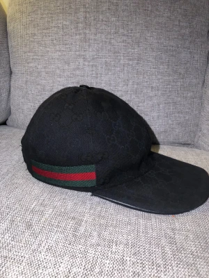 Svart Gucci keps - Snygg svart keps från Gucci med diskret GG-logomönster över hela kepsen. På sidan finns en klassisk grön och röd rand. Justerbar rem baktill för perfekt passform. Materialet är slitstark bomull och kepsen har en böjd skärm.