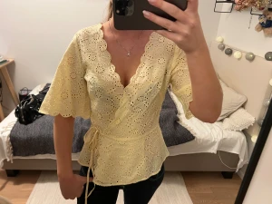 Gul omlottopp med broderade detaljer - Söt gul omlottopp med broderade hålmönster och scallop-kant. Toppen har knyt i midjan, v-ringning och korta, lite utsvängda ärmar. Perfekt för vår och sommar, ger en romantisk vibe och är superfin till både jeans och kjol.