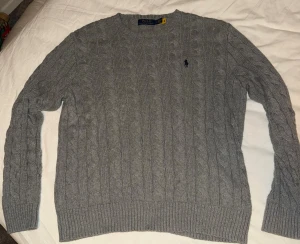 Grå stickad tröja från Polo Ralph Lauren - Snygg grå stickad tröja från Polo Ralph Lauren med klassiskt kabelstickat mönster och rund hals. Tröjan har långärmade ribbade muddar och den ikoniska Polo-loggan broderad på bröstet. Perfekt för dig som gillar stilren och tidlös design.