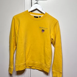 Gul sweatshirt från GANT - Snygg gul sweatshirt från GANT med broderad logga och emblem på bröstet. Tröjan har rund hals, ribbade muddar och är långärmad. Perfekt för dig som gillar klassisk stil med en färgklick. Materialet är mjuk bomull som känns skönt mot huden.