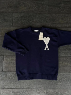 Mörkblå stickad tröja från Ami Paris - Snygg mörkblå stickad tröja från Ami Paris med vit hjärtformad A-logga på bröstet. Tröjan har rund hals, ribbade muddar och lång ärm. Perfekt för dig som gillar stilrena detaljer och vill ha något som sticker ut lite extra.