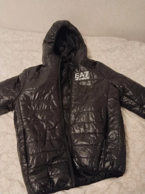Svart pufferjacka från Emporio Armani - Säljer en svart pufferjacka från Emporio Armani med huva och snyggt EA7-tryck på bröstet. Jackan har quiltad design, dragkedja framtill och är tillverkad i glansig syntet. Perfekt för dig som vill ha en stilren och varm jacka med streetkänsla.