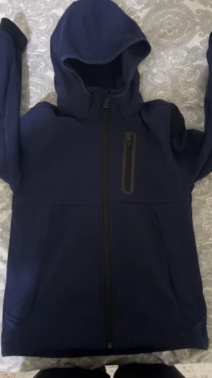 Mörkblå vindjacka från McKinley - Snygg mörkblå vindjacka från McKinley med huva och hel dragkedja framtill. Jackan har en praktisk bröstficka med dragkedja och diskret logga på ärmen. Perfekt för blåsiga dagar och outdoor-aktiviteter. Materialet är slitstark syntet som står emot vind.