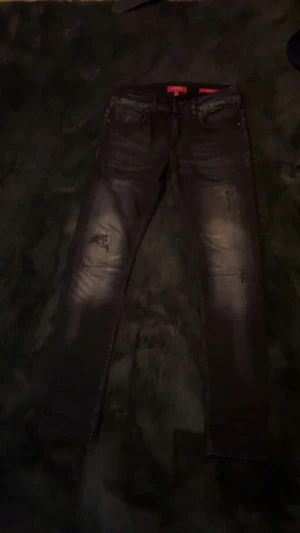 Guess svarta skinny jeans, stl 28 - Svarta skinny jeans från Guess med coola slitningar och tvättade detaljer. Klassisk femficksmodell med smal passform och låg midja. Snygga kontrastsömmar och Guess-logga på bakfickan. Perfekta för dig som gillar en edgy stil.