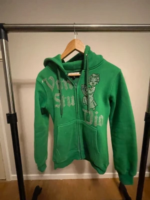 Hoodie med glittertryck från Vonstudio  - Cool grön hoodie från Vonstudio i storlek XS med glittrigt tryck och motiv framtill. Tröjan har huva med snörning, dragkedja och två fickor. Perfekt för dig som gillar streetstyle och vill sticka ut med färg och detaljer.