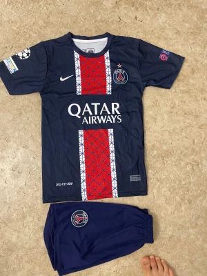 PSG fotbollströja D.Doué #14 Nike - Paris Saint-Germain fotbollströja med D.Doué och nummer 14 på ryggen. Mörkblå med röd och vit randig panel framtill, PSG-logga och Qatar Airways tryck. Kortärmad, tillverkad i lätt polyester med Dri-Fit ADV. Matchande mörkblå shorts med PSG-emblem ingår.