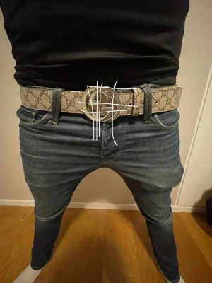 Beige Märkes Bälte - Säljer ett snyggt beige bälte med GG-mönster och stor rund silverspänne. Bältet är gjort i skinn och har en lyxig vibe som lyfter din outfit. Perfekt för dig som vill sticka ut med ett statement piece från ett ikoniskt märke.