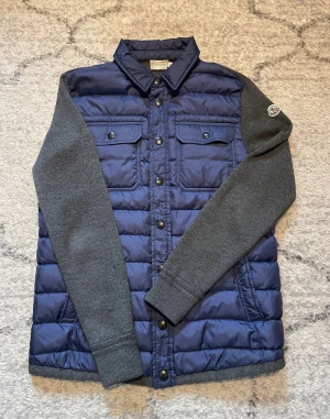 Blå och grå pufferjacka från Moncler - Säljer en snygg cardigan från Moncler med blå, quiltad framsida och grå stickade ärmar och rygg. Jackan har två bröstfickor med knappar, sidofickor och Moncler-logga på ärmen. Perfekt för dig som gillar streetstyle och vill ha något unikt.