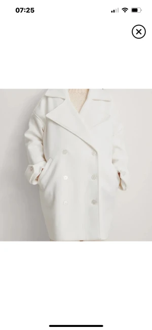 Vit oversized kappa med knappar - Snygg vit kappa i oversized modell med breda slag och dubbelknäppning framtill. Jackan har långa ärmar, fickor på sidorna och dekorativa knappar vid ärmslut. Perfekt för dig som gillar en clean och stilren look.