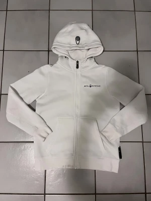 Vit zip hoodie från Sail Racing bowman strl 160 - Vit zip hoodie från Sail Racing bowman pojke strl 160 med dragkedja och huva. Tröjan har logga på bröstet och ett tryck på huvan. Praktiska fickor framtill och ribbade muddar vid ärmslut och nederkant. Perfekt för chill dagar eller när du vill ha en clean look. Tröjan har en fläck nere vid kanten av tröjan. 