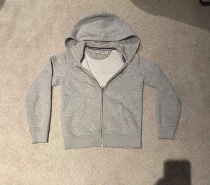 Grå hoodie från GANT med dragkedja - Snygg grå hoodie från GANT med hel dragkedja framtill och huva. Den har även två sidofickor. Insidan av kragen har en cool randig detalj i rött, vitt och blått. Perfekt för chill dagar och enkel att matcha.