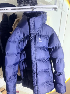 Blå pufferjacka från Haglöfs - Säljer en blå pufferjacka från Haglöfs i storlek XL Jackan har hög krage, markerade sömmar och Haglöfs-logga på bröstet. Materialet är syntet och dun, perfekt för kalla dagar. Jackan har lång ärm och dragkedja framtill.