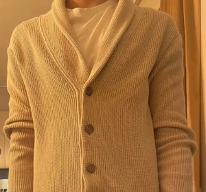 Beige kofta från John Henric - Säljer en beige stickad kofta från John Henric i storlek S. Koftan har ribbad struktur, bred krage och knäpps med fem bruna knappar framtill. Perfekt för lager-på-lager och chill stil under höst och vinter.