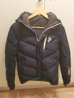 Mörkblå Peak Performance dunjacka 160 - Snygg mörkblå dunjacka från Peak Performance i juniorstorlek 160. Jackan har huva, dragkedjor i kontrastfärg, logga på bröstet och flera fickor. Perfekt för kalla dagar och riktigt skön att ha på sig.