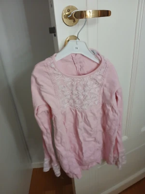 Rosa blus med spets från Sweet Me - Supersöt ljusrosa blus från Sweet Me i storlek 122/128. Blusen har långa ärmar och vacker spets med blommigt broderi framtill samt spetsdetaljer vid ärmslut. Rund halsringning och knapp i nacken. Perfekt för dig som gillar romantisk stil!