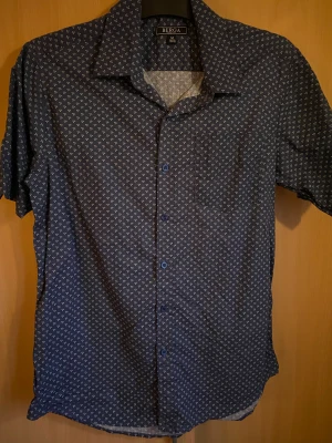Mörkblå mönstrad skjorta från Berga - Snygg mörkblå skjorta från Berga med små vita mönster över hela tyget. Skjortan har korta ärmar, klassisk krage och knappar framtill. Perfekt för dig som gillar stilrena detaljer och vill ha en lätt skjorta i bomull.