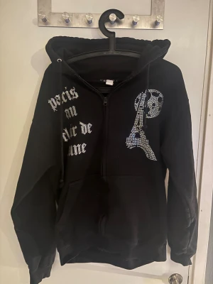 Svart hoodie med strass och Paris-tryck - Svart hoodie från Blinglucid med strassdetaljer som bildar texten 'paris au clair de lune' och motiv av Eiffeltornet och månen. Tröjan har huva med snörning, dragkedja framtill och stora fickor. Perfekt för dig som gillar statement-plagg och coola detaljer. Dragkädjan går inte att dra ner hela vägen.
