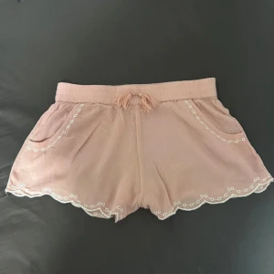 Rosa shorts med broderad spetskant - Söta ljusrosa shorts med snörning i midjan och vita broderade detaljer längs fickor och vågig spetskant. Luftigt material och avslappnad passform
