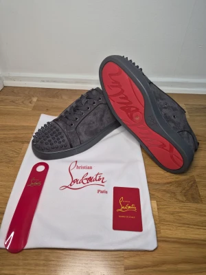 Grå Christian Louboutin sneakers med nitar - Snygga grå sneakers från Christian Louboutin i mocka med ikonisk röd sula och coola nitar på tån. Skorna har snörning och en rund tå, samt en platt sula. Perfekt för dig som vill sticka ut med exklusiv design och edgy detaljer.