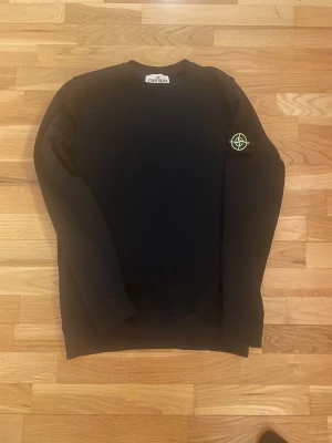 Svart sweatshirt från Stone Island - Svart sweatshirt från Stone Island med klassisk rund hals och ikonisk logotyp-patch på vänster ärm. Tröjan har lång ärm och en clean, stilren look som passar perfekt till streetwear. Materialet känns mjukt och bekvämt, troligtvis bomull eller polyester.