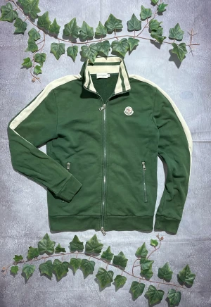 Moncler Grön Sweatshirt Storlek S/M [ÄKTA] - 🎨 Färg: Grön med vita ränder längs ärmarna 📏 Storlek: S/M ♻️ Skick: 9/10 – lättare bruksspår men inga större defekter 🧵 Material: Bomullsblandat sweatshirt-tyg  ✨ Detaljer:         •        Hög krage         •        Tvåvägsdragkedja framtill         •        Två framfickor med dragkedja         •        Moncler-patch på bröstet         •        Ribbstickade muddar och midja    