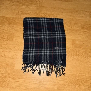 Rutig halsduk från Burberry  - Snygg lång halsduk från burberry med klassiskt rutigt mönster i mörkblått, grått och röda linjer. Gjord i cashmere och har fransar i ändarna. Perfekt att slänga över axlarna för en cool och tidlös look.