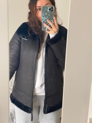 Svart pälsjacka med dragkedja - Säljer en svart pälsjacka med fluffig krage och muddar. Jackan har snygga detaljer som spänne vid axeln och dragkedjor på fickorna. Perfekt för dig som vill ha en varm och stilren look under kalla dagar. Jackan är lite längre i modellen och har en skön, loose passform.