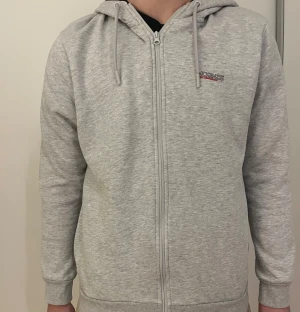 Ljusgrå hoodie från McKenzie - Säljer en ljusgrå hoodie från McKenzie med dragkedja och huva. Tröjan har snörning i huvan, ribbade muddar och en liten logga på bröstet. Perfekt för chill dagar eller när du vill ha en enkel och clean look.