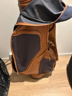 Cobra golfbag i blått tyg och brunt skinn - Snygg golfbag från Cobra i mörkblå textil med bruna skinnpaneler och detaljer. Väskan har flera dragkedjeförsedda fack, broderad logga och vadderad axelrem. Perfekt för dig som vill ha en stilren och klassisk look på golfbanan. Det får plats med många golfklubbor och massa förbackning som extra kläder, mat, dryck och mer.