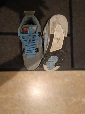 Nike Air Jordan 4 University Blue - Nike Air Jordan 4 University Blue sneakers med ljusblå mocka, vita och svarta detaljer samt meshpaneler på sidorna. Skorna har snörning, Jumpman-logga på hälen och platt sula med blå och svarta inslag. Perfekt för dig som gillar streetwear och klassiska Jordan-vibbar.