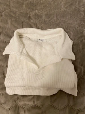 Vit ribbad pikétröja från Kappahl - Vit pikétröja från Kappahl i ribbad bomull med klassisk krage och korta ärmar. Tröjan har en enkel och clean look, perfekt för dig som gillar stilrena plagg. Materialet är mjukt och har en snygg struktur som ger lite extra känsla.