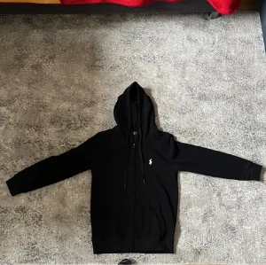Svart hoodie från Polo Ralph Lauren - Svart hoodie från Polo Ralph Lauren med klassisk vit broderad logga på bröstet. Tröjan har huva med snörning, dragkedja framtill och långa ärmar. Mjuk bomullskvalitet som är skön att bära. Perfekt för dig som gillar stilrena och tidlösa plagg. Mindre än S skulle jag säga. 